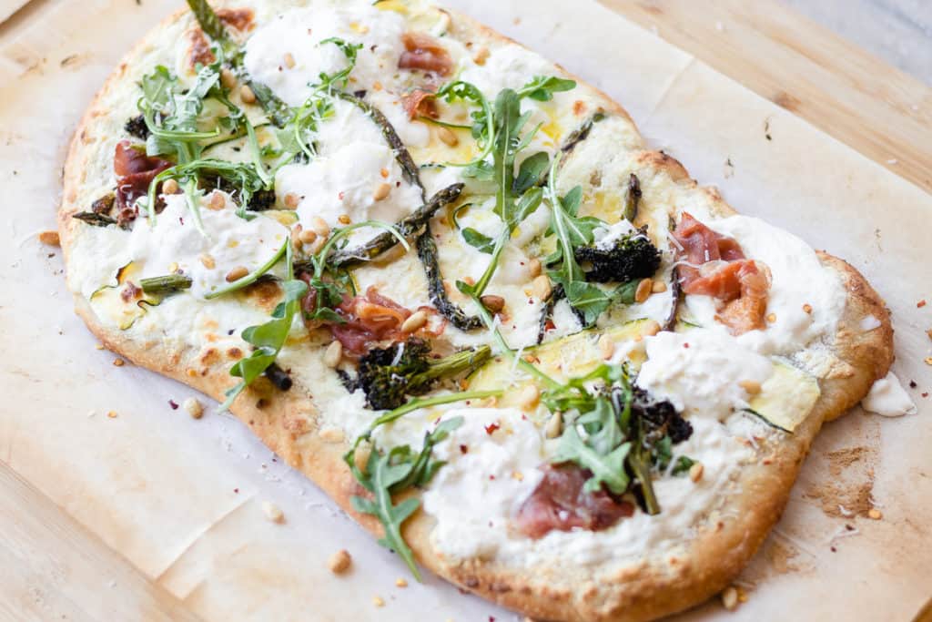 veggie pizza on parchment paper with white sauce and pine nuts