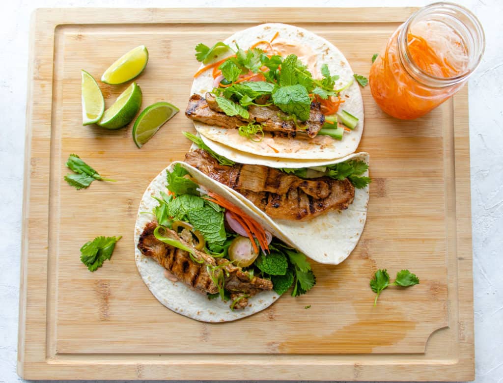 Tacos on a cutting board, filled with grilled meat, pickled veggies and herbs
