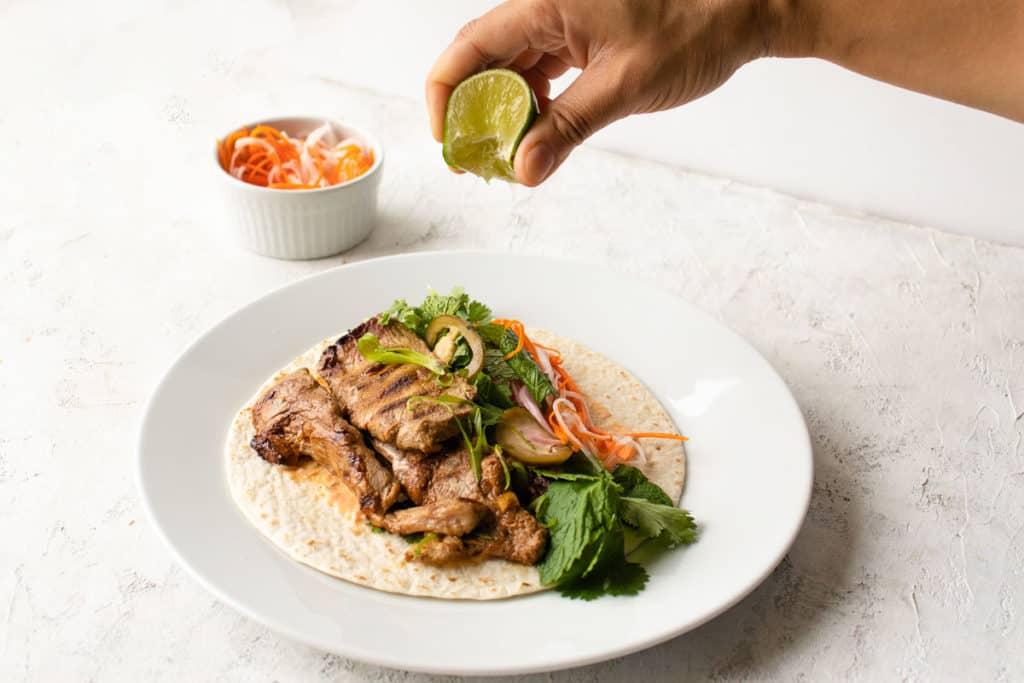 A lime being squeezed over bahn mi pork tacos with herbs, jalapenos and pickled vegetables