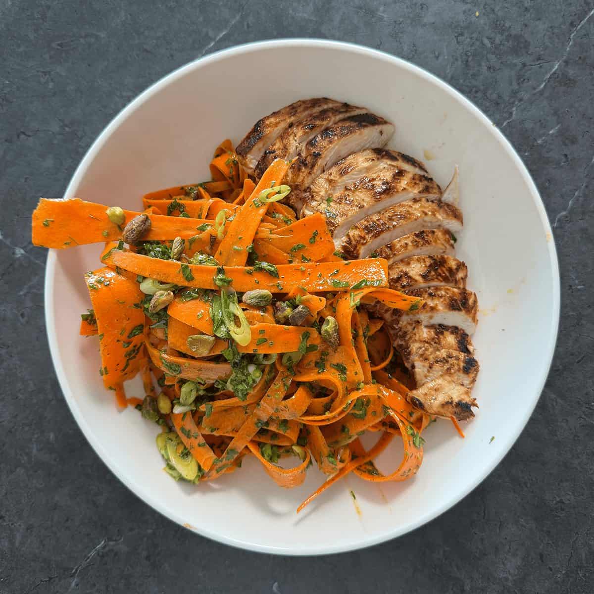 Grilled chicken over carrot ribbon salad plated in a white bowl