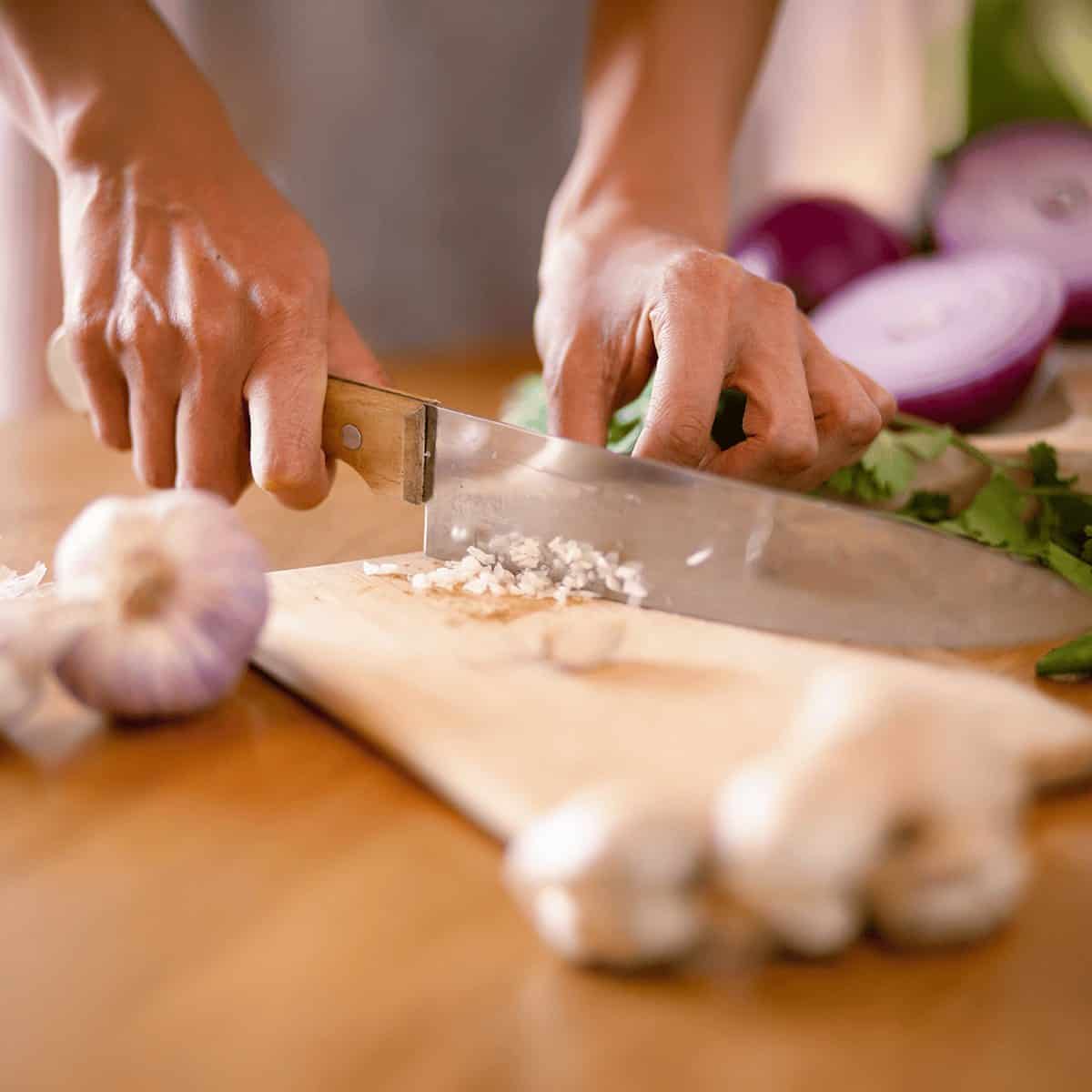 A chef’s hands mincing garlic on a wooden cutting board