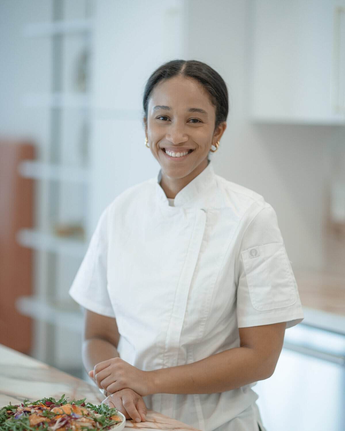 Eats by Nicole founder Nicole Lee in chef whites at a kitchen counter with a plated salad